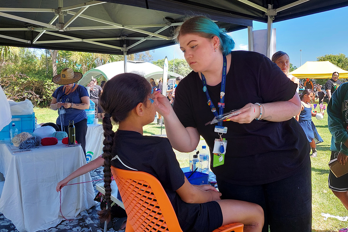 A woman with blue hair doing face painted on a girl seated with her back to the camera.